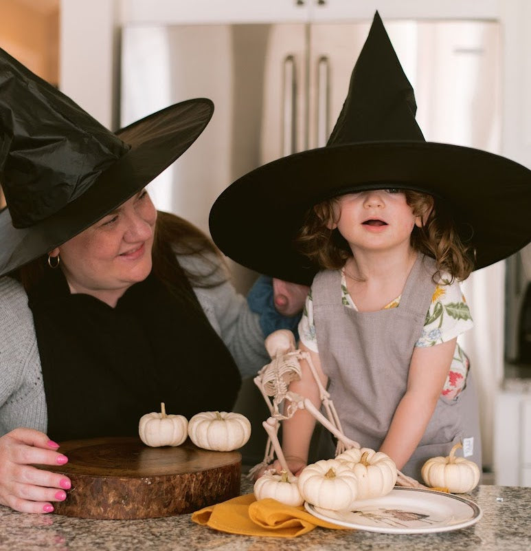 A mom and a little girl wearing aprons and witch hats play with skeletons and pumpkins